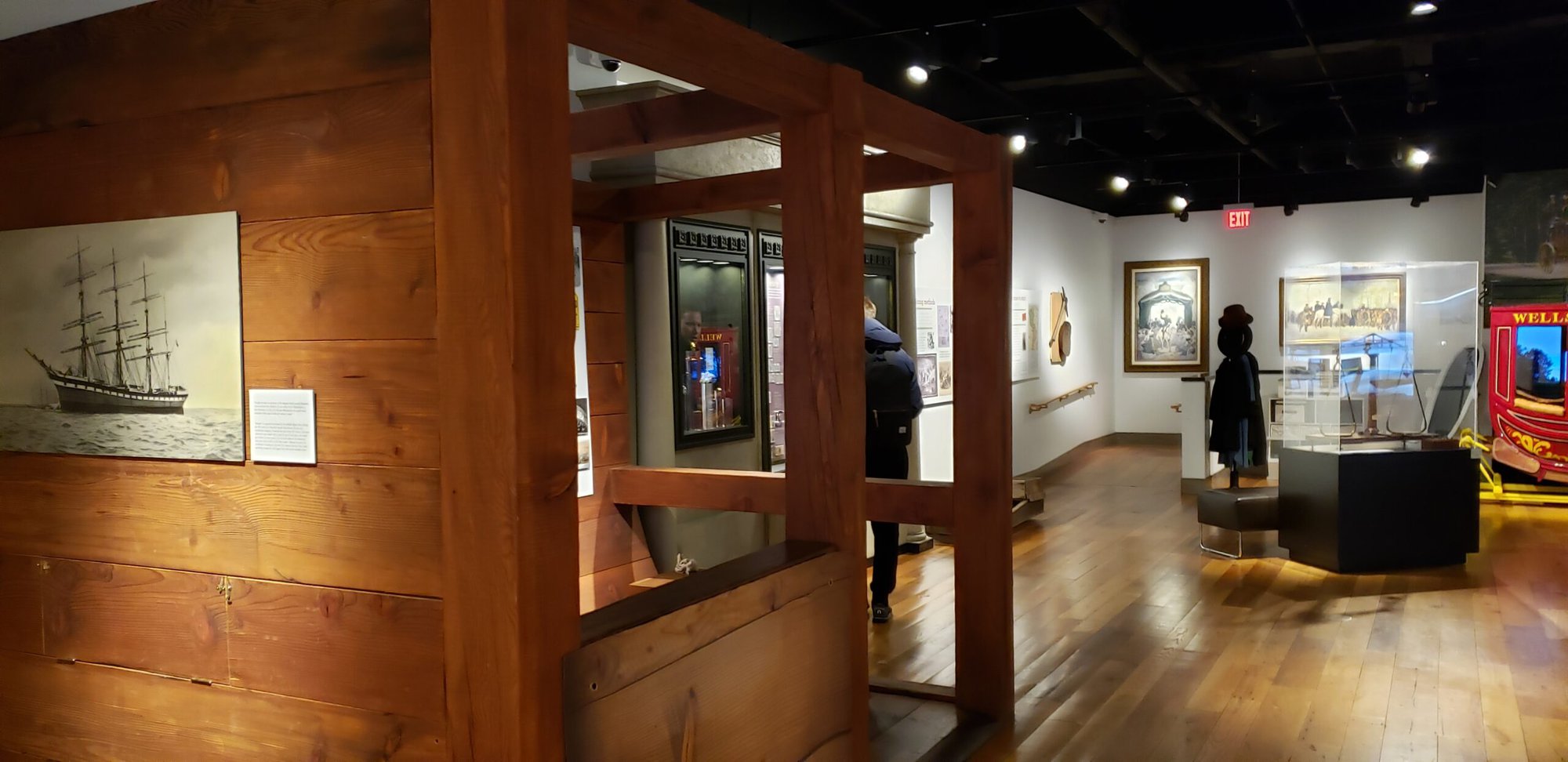 Interior of a museum exhibit featuring wooden structures, historical artifacts, paintings, and display cases. A person is viewing an exhibit in the background, and a model ship painting is visible on a wooden wall in the foreground.