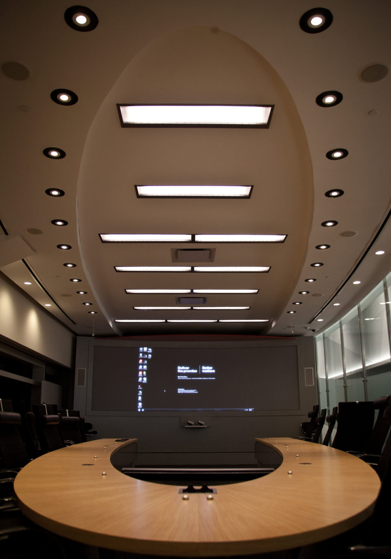 Modern conference room with a large curved wooden table, ergonomic chairs, and a ceiling with recessed lighting and rectangular light fixtures. A large screen displays a computer desktop with the text 'Deliver the promise' and 'Better matters.